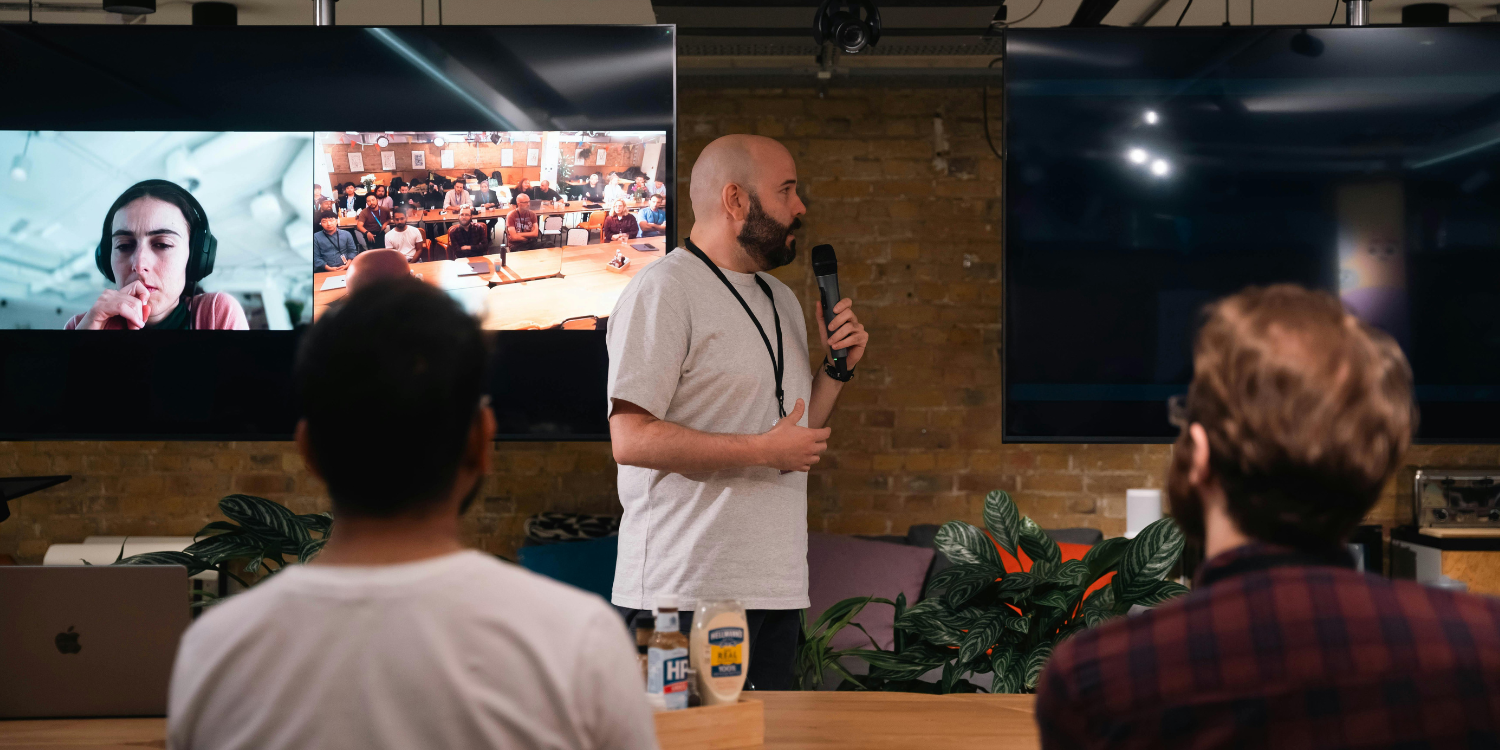 Man speaking at an event, screens show a video call.