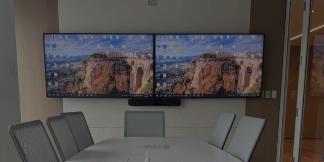 Meeting room with two large screens displaying a desktop image, table with chairs.