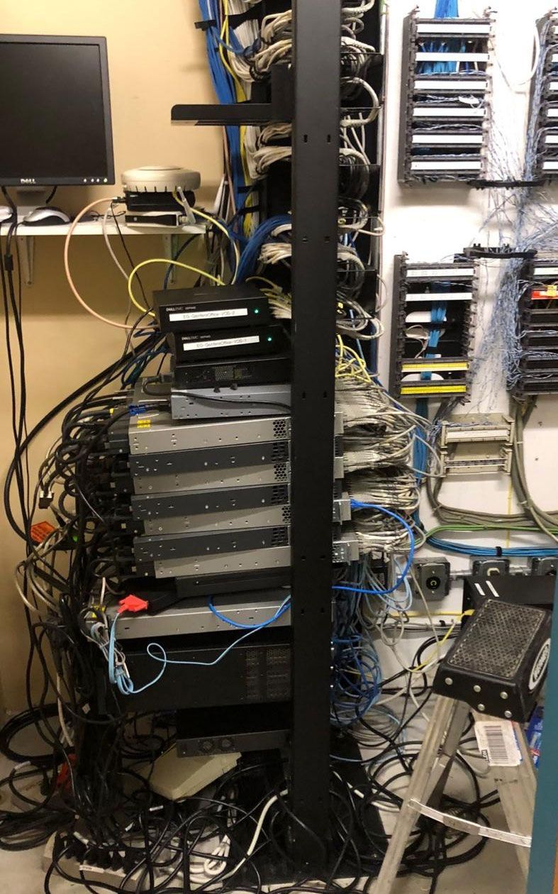 Server rack with tangled cables, surrounded by wall-mounted network hardware and a monitor.