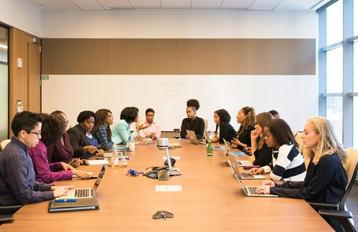 A diverse group of people sitting around a long conference table, working on laptops in a brightly lit office.