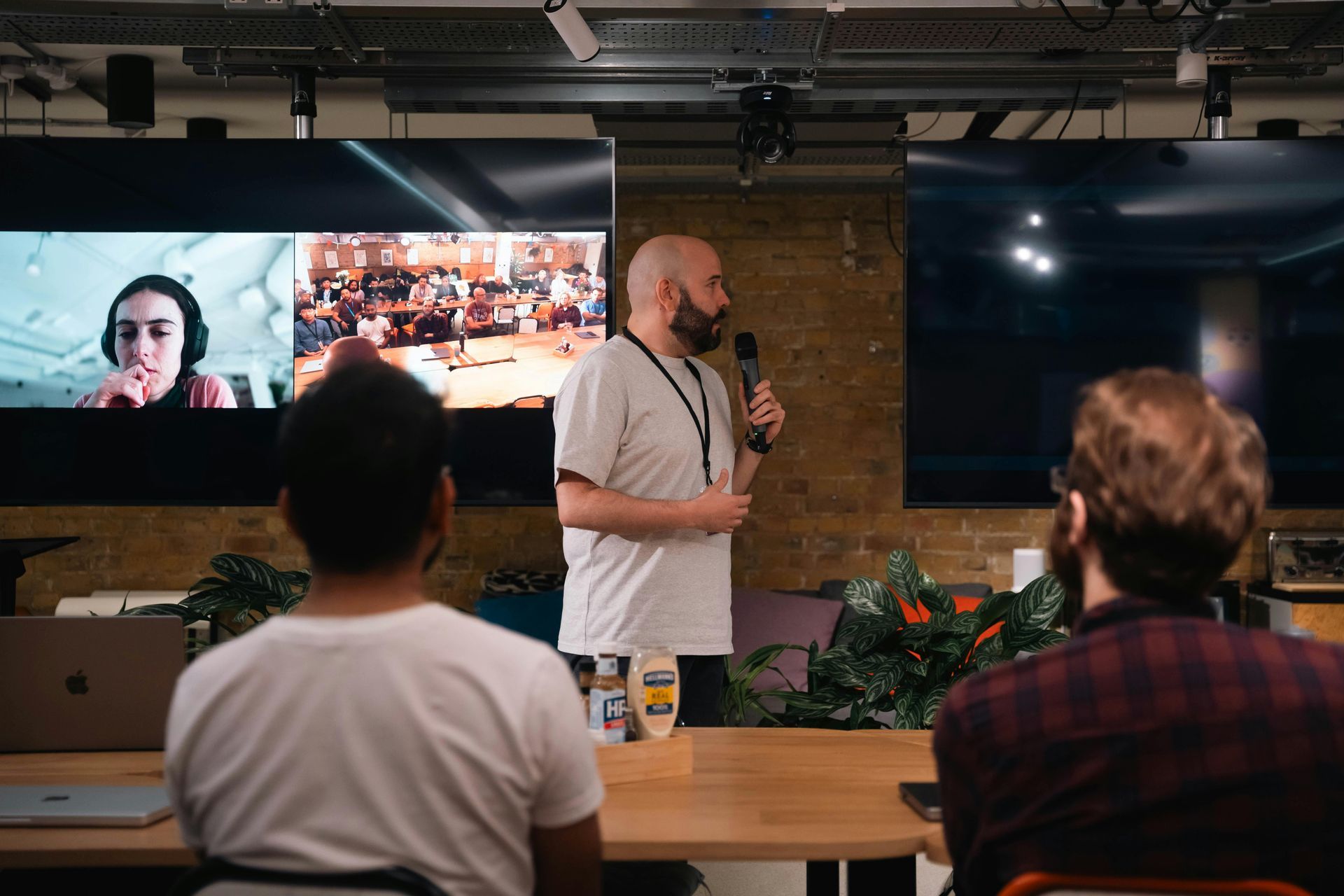 Man speaking at a presentation, screens showing remote participants.