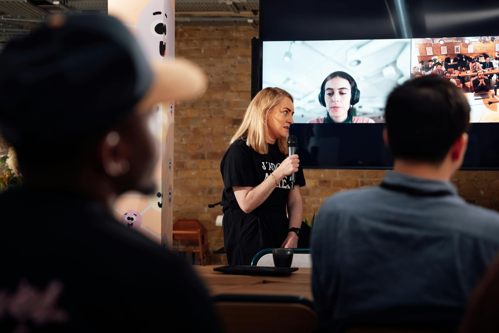 A woman with a microphone speaks at an event while a video call plays on a screen behind her.