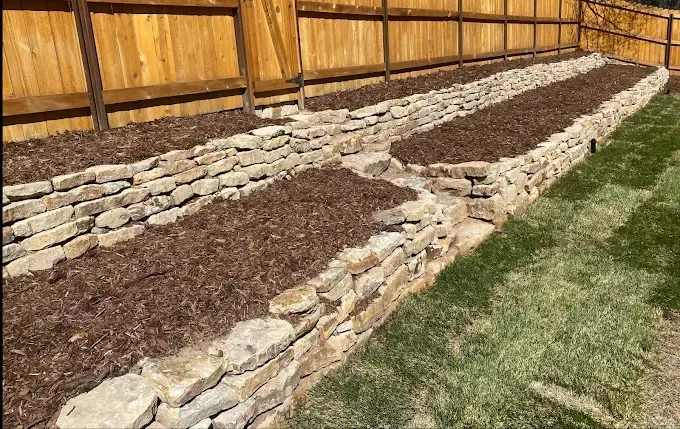 Stone-walled raised garden beds filled with mulch, next to a wooden fence and grass lawn.