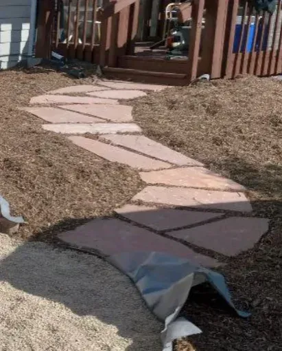 Stone pathway through wood-chipped landscaping, leading to wooden deck with steps. Grey fabric visible in the foreground.