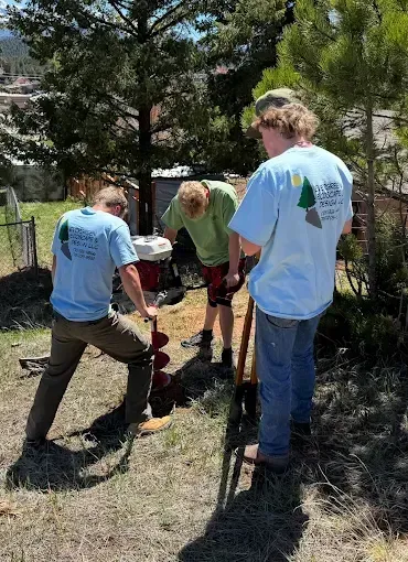 Three people drilling into the ground outdoors, wearing light blue and green shirts.