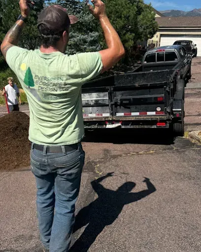Man with arms raised, sweaty, stands before a trailer. Another person works near a pile of mulch. Sunny day.