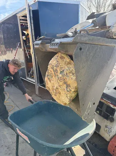 Man uses a machine to load a large rock into a wheelbarrow near a trailer.