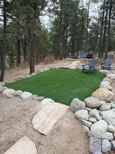 A small, green lawn surrounded by rocks, with Adirondack chairs in a wooded setting.