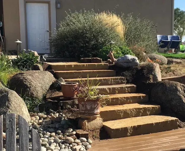 Stone steps leading up to a white door, surrounded by plants, rocks, and a wooden walkway.