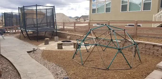 Backyard with a trampoline, climbing dome, fire pit, and a house. Brown mulch and a concrete path.