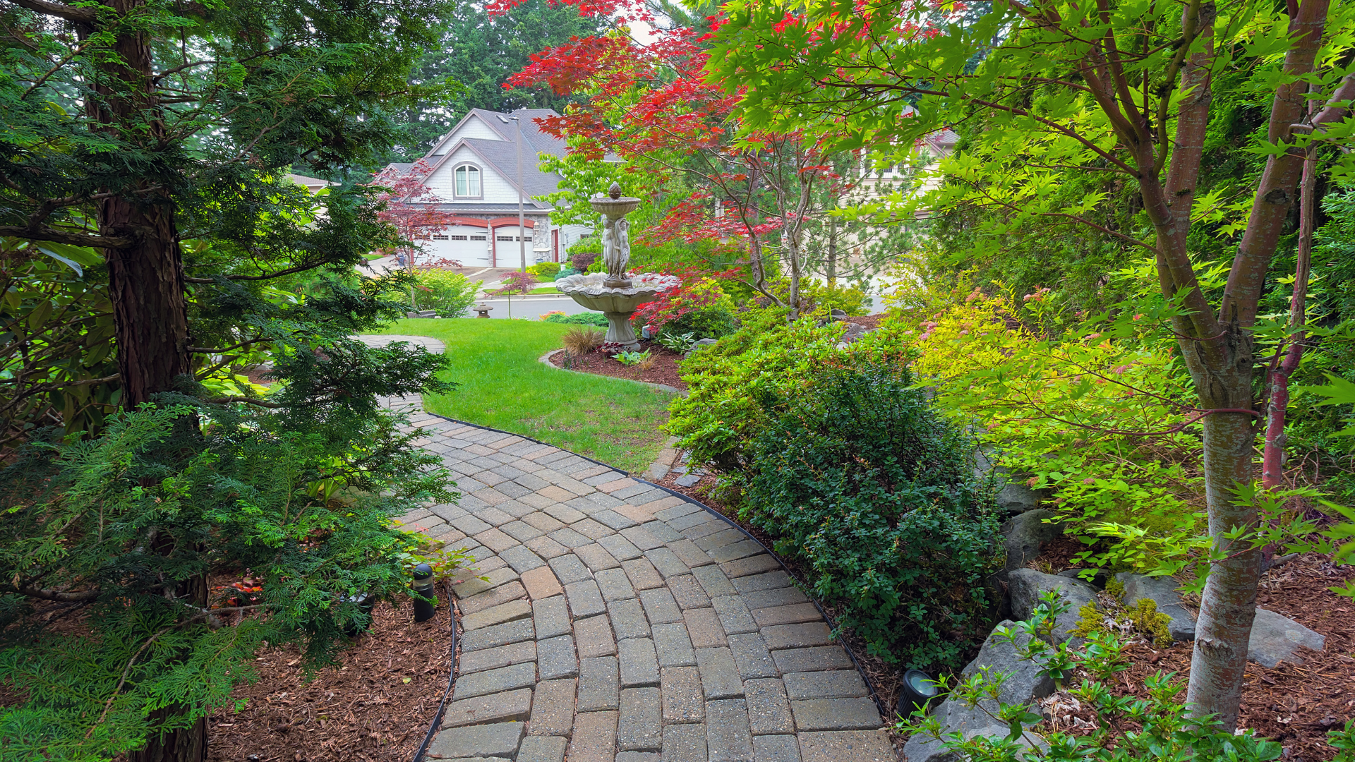 A stone path winds through a lush, green garden toward a fountain, with a house visible in the background.