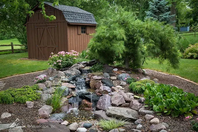 A brown shed sits behind a small waterfall and rock garden, surrounded by greenery.