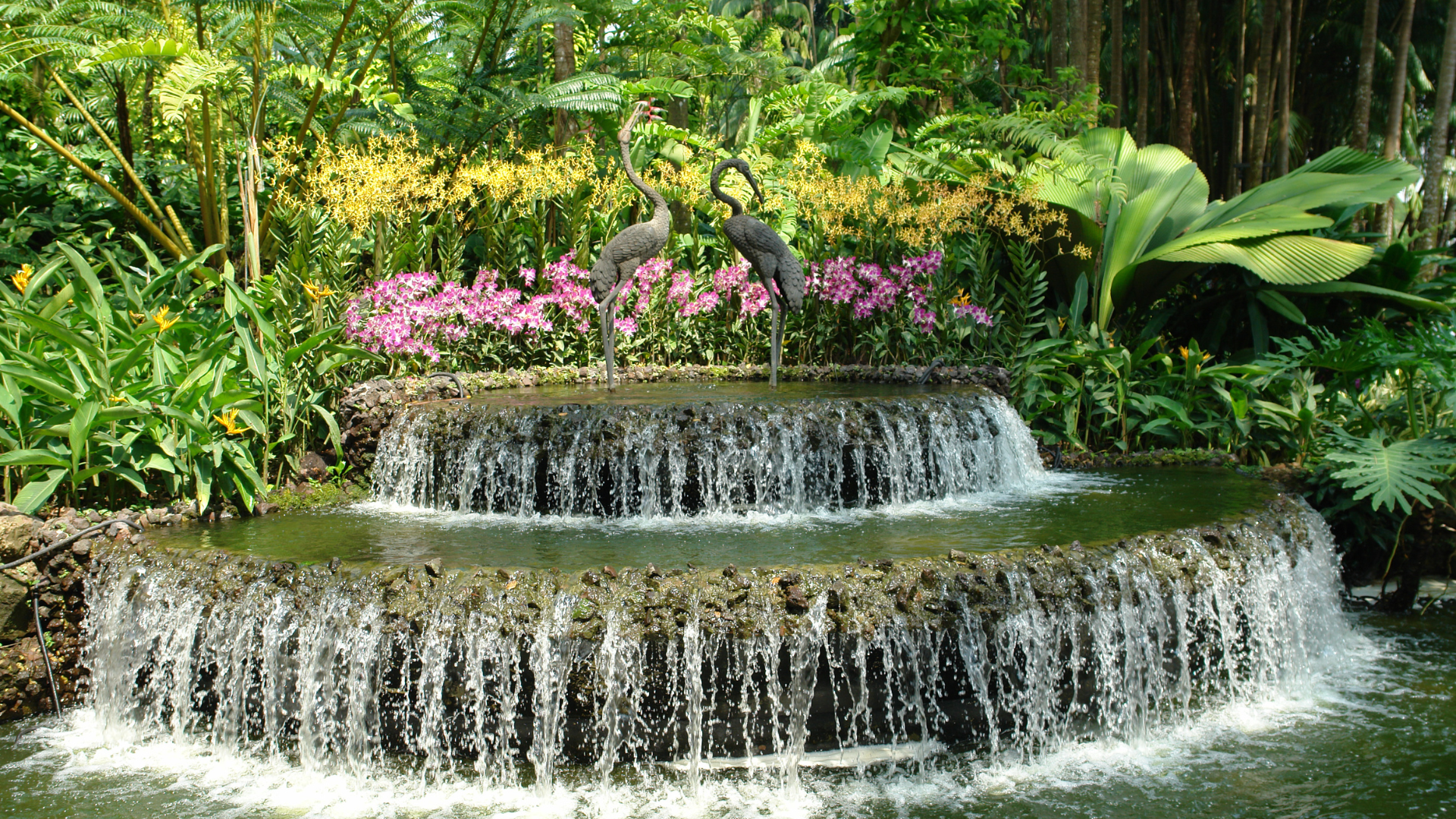 A two-tiered tiered stone fountain with water cascading into a pool, surrounded by lush greenery and pink orchids.