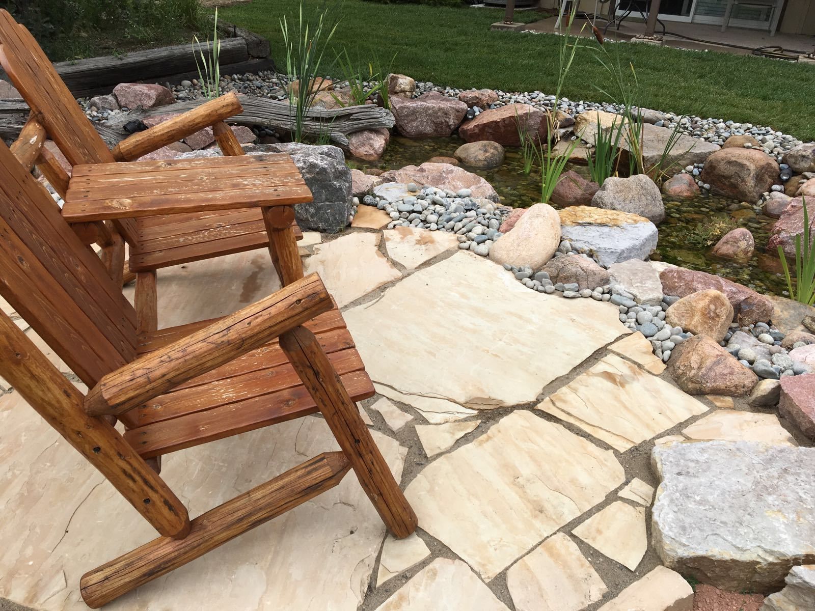 Wooden Adirondack chair on stone patio next to a rock-lined pond.