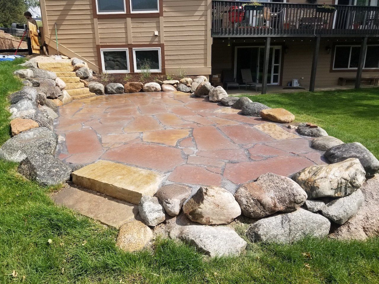 Stone patio with steps, surrounded by rocks and green grass, next to a house.
