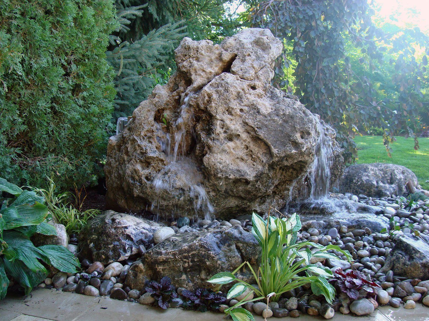Water fountain made of large rock, water cascading down, surrounded by plants and pebbles.