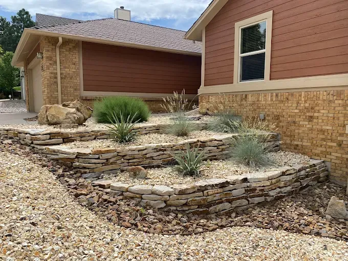 Stone tiered garden with drought-tolerant plants next to a house with tan stone and brown siding.