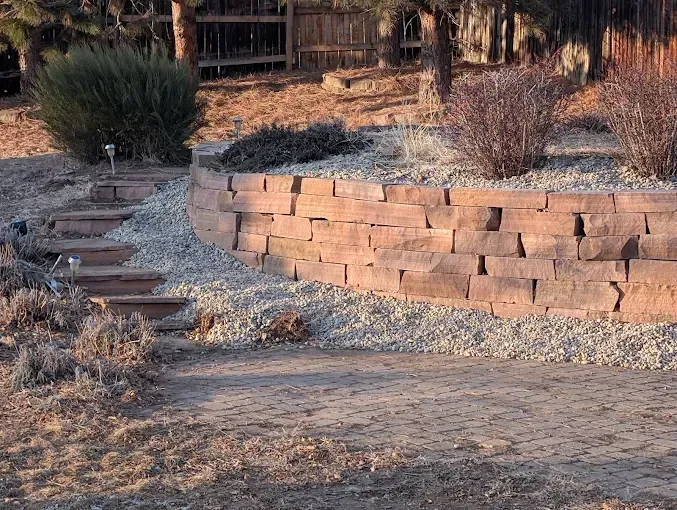 Stone retaining wall with steps leading up, gravel, and sparse landscaping in yard.