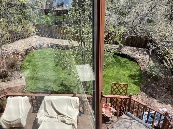 Backyard view: green lawn, wooden deck with covered chairs, brown fences, trees, and partial reflection in window.
