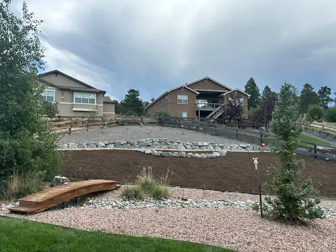 Backyard landscape with two houses, a rock garden, and a small wooden bridge under a cloudy sky.