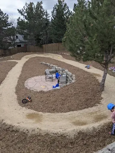 Backyard with a wood chip track for toy cars, a patio area with a table, and a child watching.