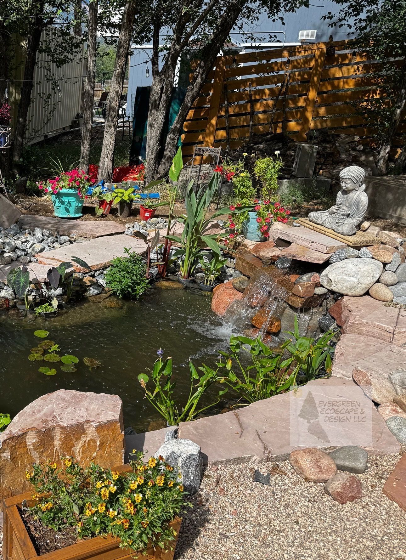 Water fountain made of large rock, water cascading down, surrounded by plants and pebbles.