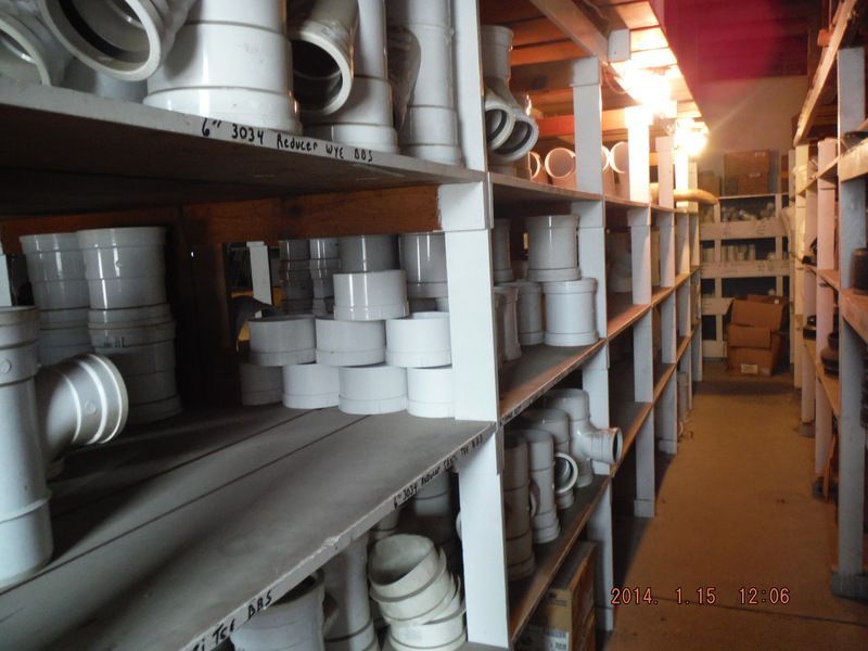 Shelves stocked with white plumbing fittings inside a dimly lit warehouse, pipes, elbows, and connectors are visible.