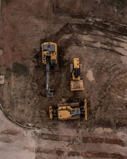 Aerial view of construction site with three yellow construction vehicles: excavator, loader, and bulldozer on muddy terrain.