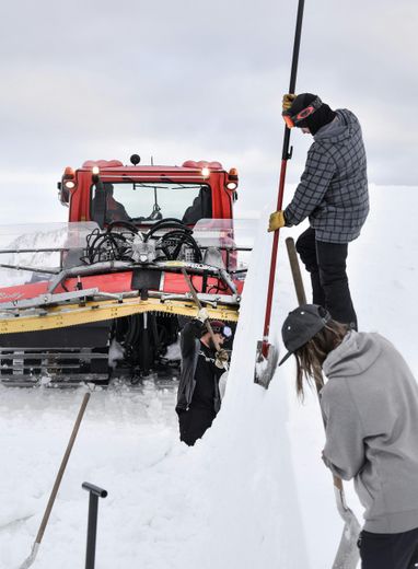 Two people shoveling snow off a large snowdrift next to a red snow groomer. Cloudy, outdoor setting.