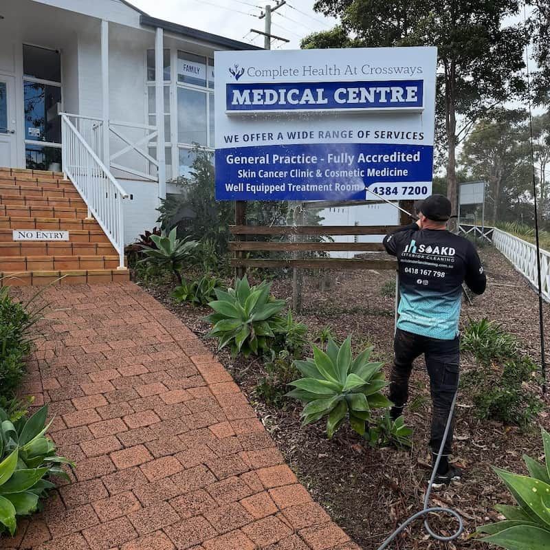 A Person in a Uniform is Cleaning a Medical Centre Sign With a Soapy Brush — SOAKD Exterior Cleaning in Newcastle, NSW