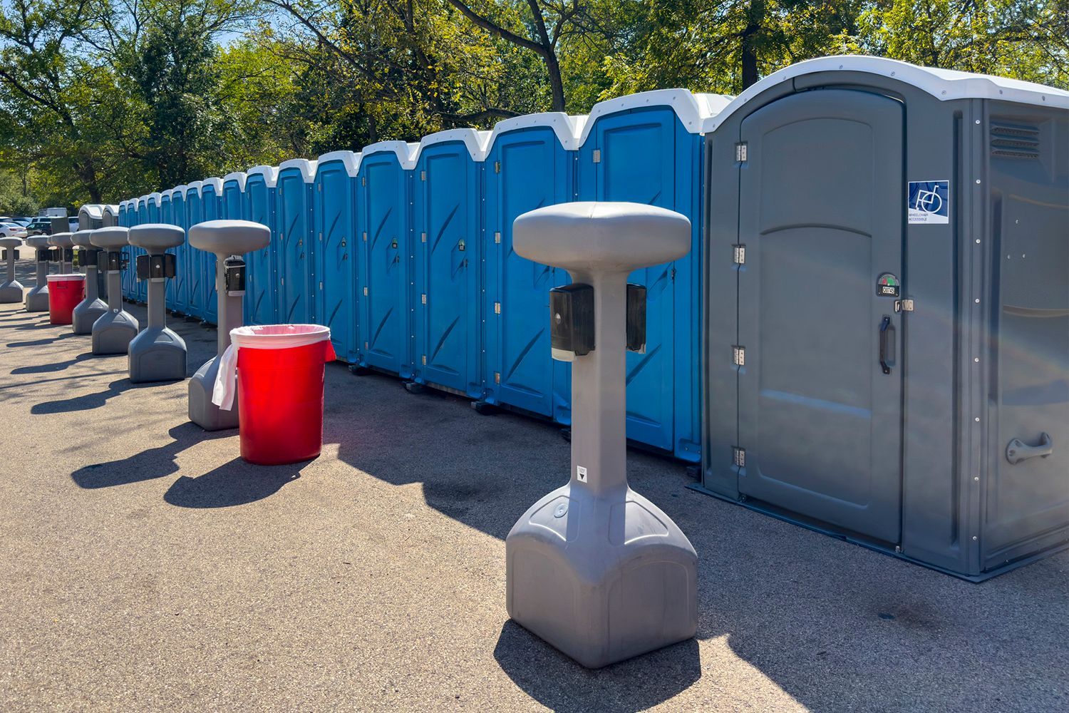 Row of blue and gray portable restrooms with hand sanitizer stations on a paved surface outdoors. Row of blue and gray portable restrooms with hand sanitizer stations on a paved surface outdoors.