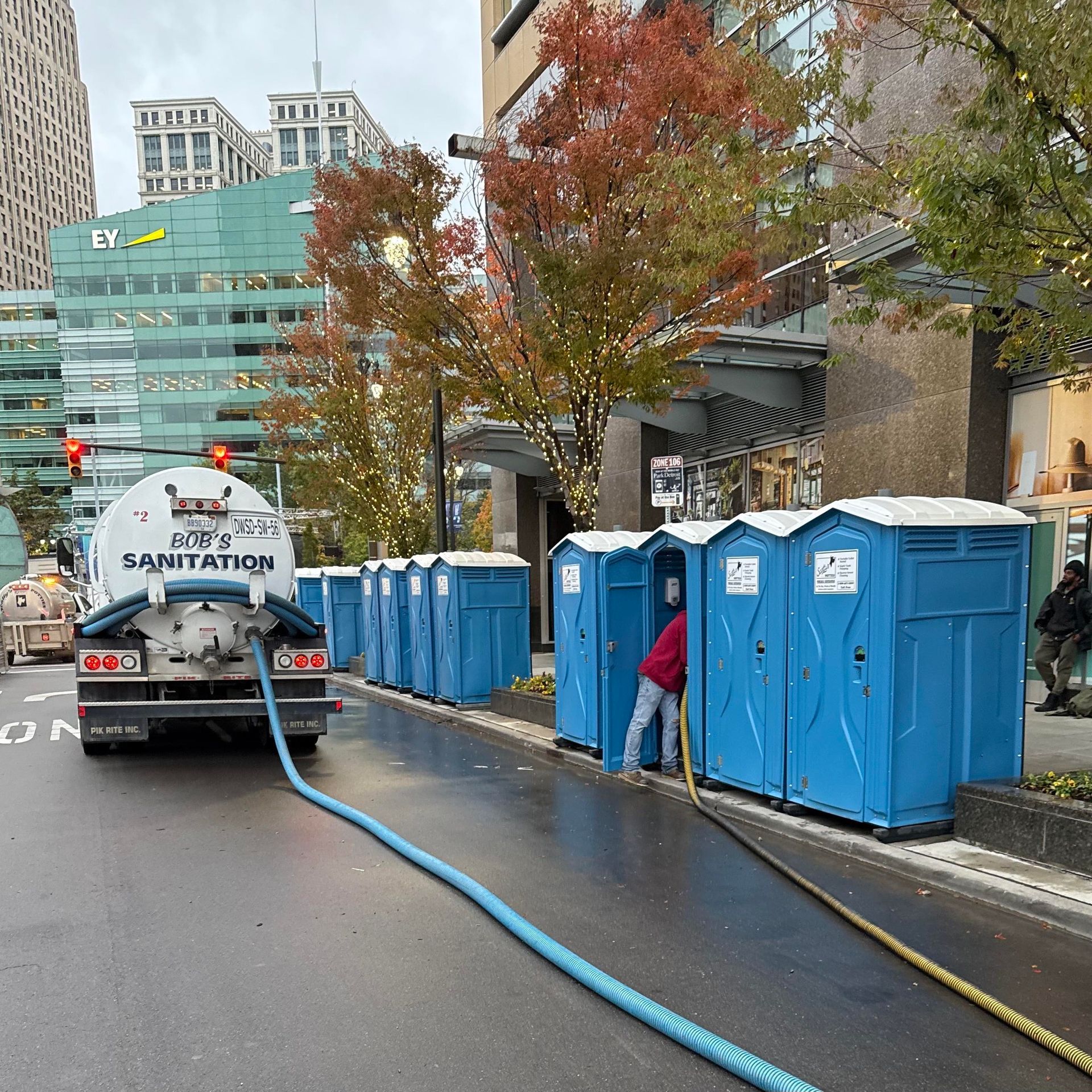 A sanitation truck servicing a row of blue portable toilets on a city street. A worker is present. A sanitation truck servicing a row of blue portable toilets on a city street. A worker is present.
