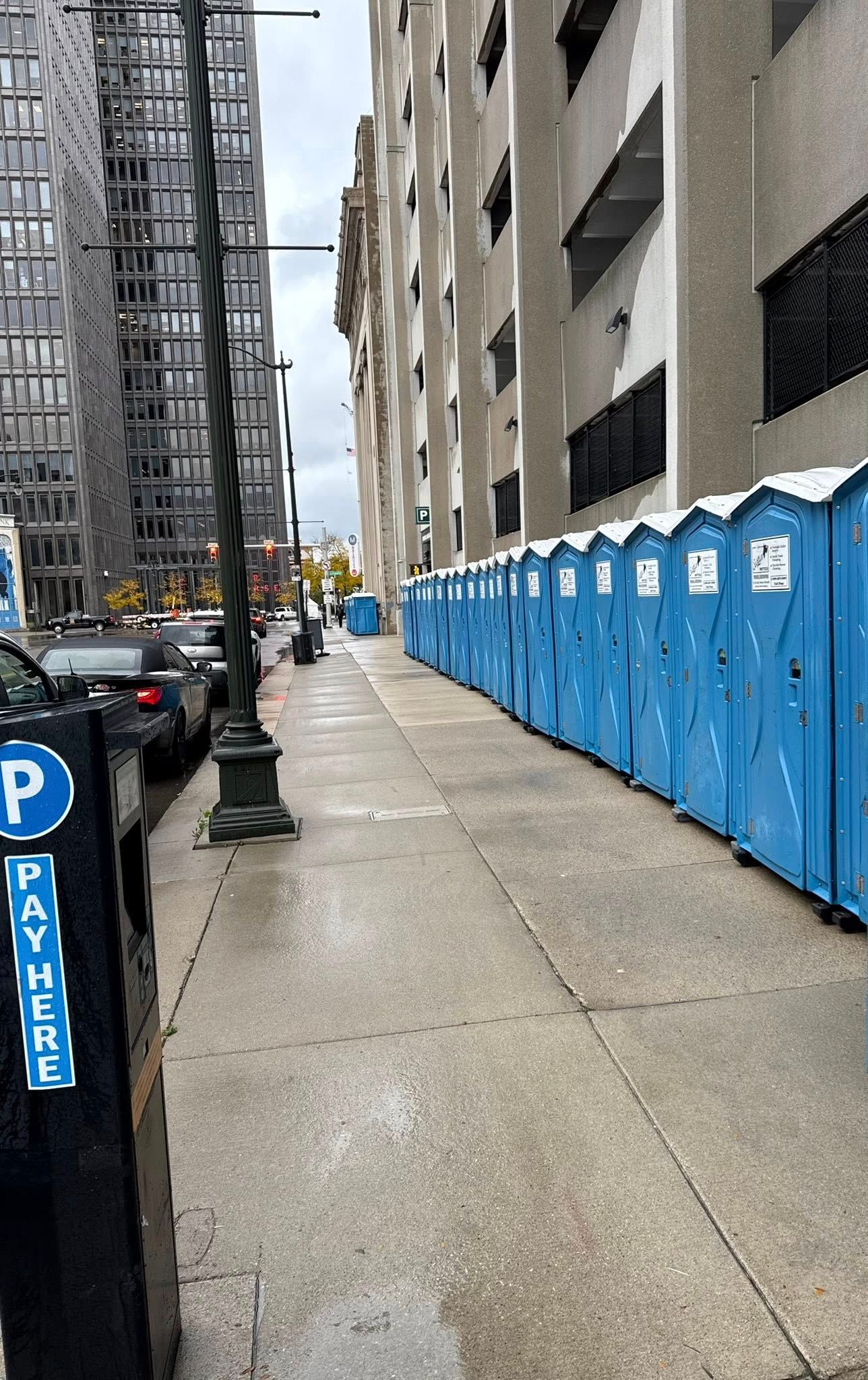 Worker in safety vest pushing a teal portable toilet off a truck bed, other toilets in the background. Worker in safety vest pushing a teal portable toilet off a truck bed, other toilets in the background.