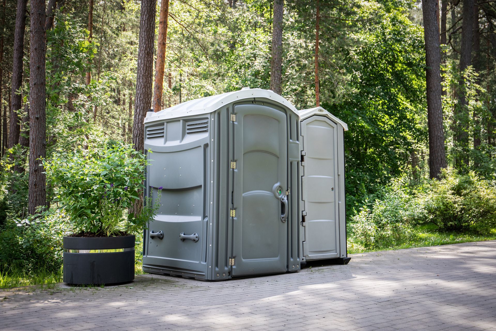 Two portable toilets on a paved path in a wooded area. One is grey, the other white. A potted plant is nearby.