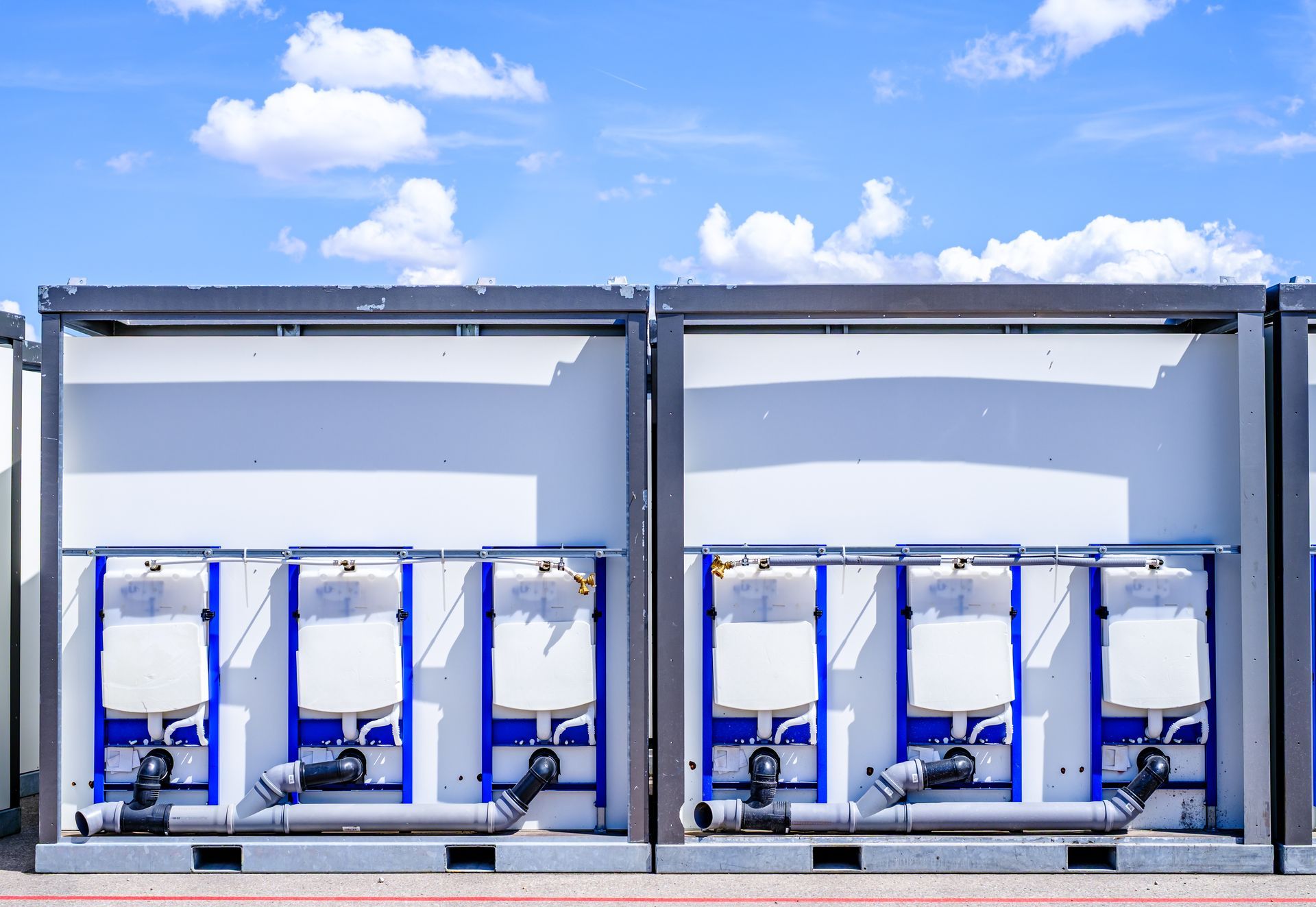 Two portable toilets with white doors, blue accents, and grey plumbing, against a bright blue sky. Two portable toilets with white doors, blue accents, and grey plumbing, against a bright blue sky.