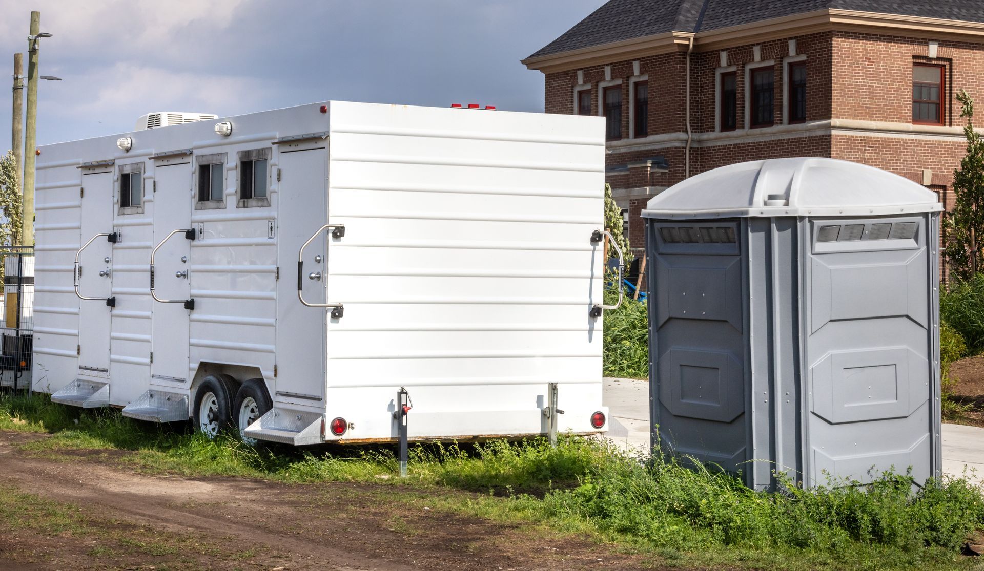 White horse trailer next to a grey portable toilet on grass near a house and road. White horse trailer next to a grey portable toilet on grass near a house and road.