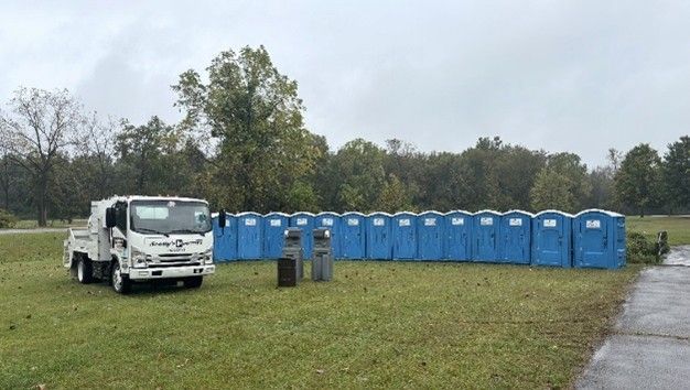 A white service truck parked on a grass field beside a long row of blue portable toilets.