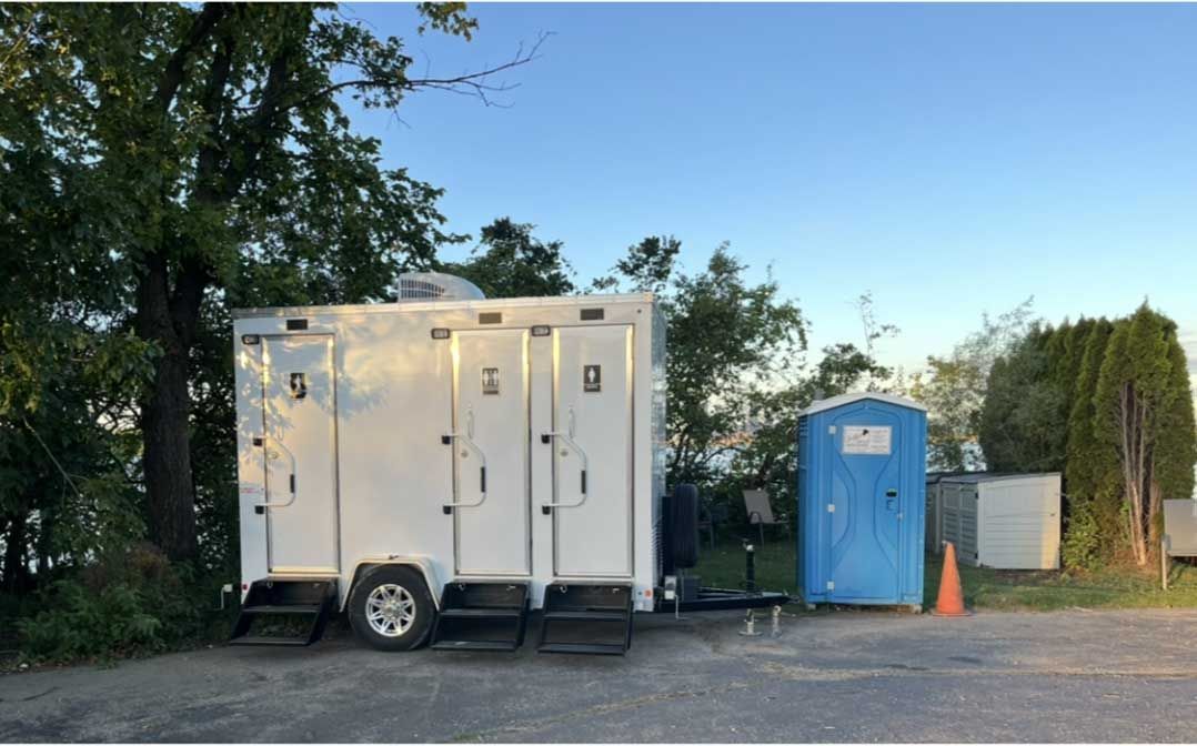 A white portable restroom trailer with three doors parked next to a single blue portable toilet on a gravel lot.