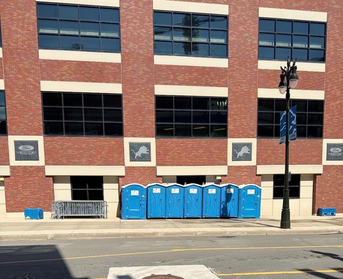Four blue portable toilets with red and white trim, lined up in a grassy outdoor area. Four blue portable toilets with red and white trim, lined up in a grassy outdoor area.