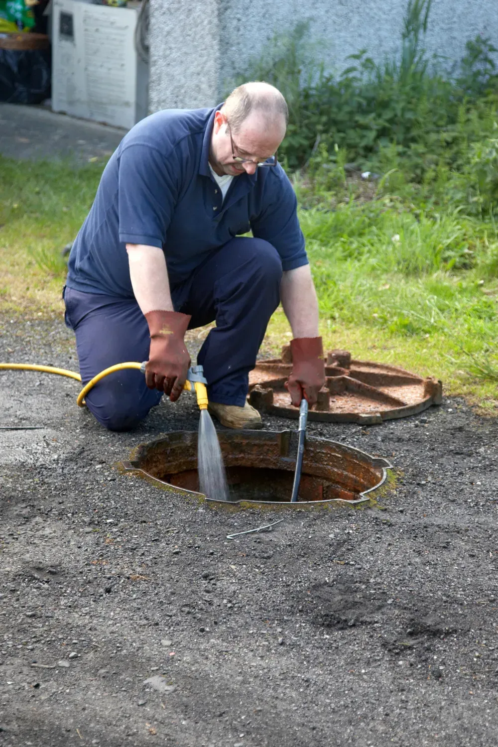 Man kneeling, cleaning a manhole with a hose. Outdoors.