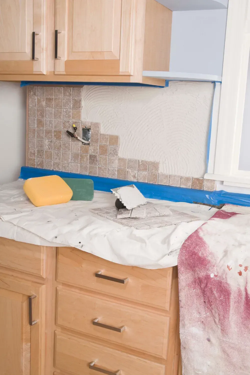 Kitchen with partial tile backsplash installation: beige cabinets, tan tile, tools, sponges, blue tape.
