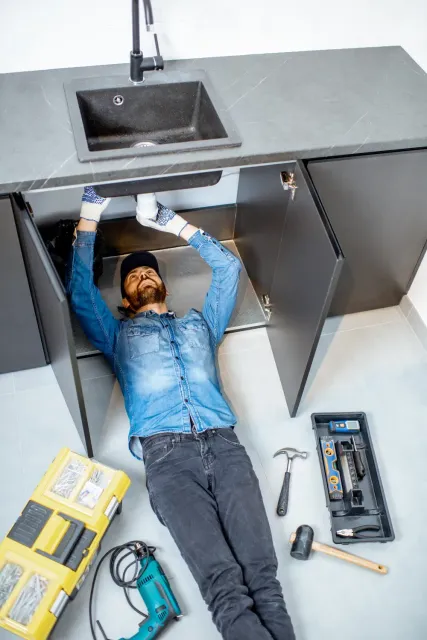 Plumber in blue shirt and jeans works under a kitchen sink with tools nearby.