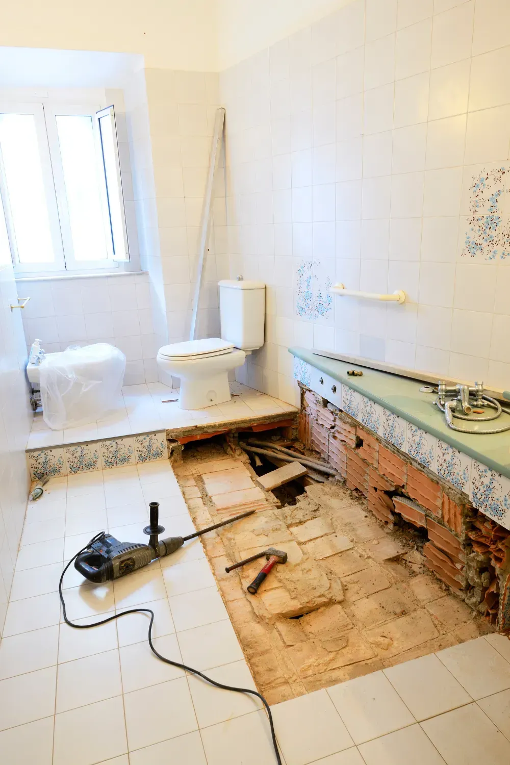 Bathroom renovation: floor tiles removed, toilet, brick wall exposed, power drill, hammer, and debris.