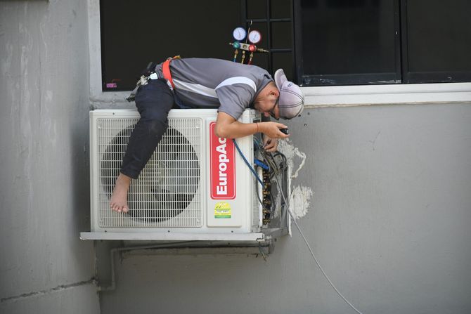 A man is laying on top of an air conditioner on the side of a building.