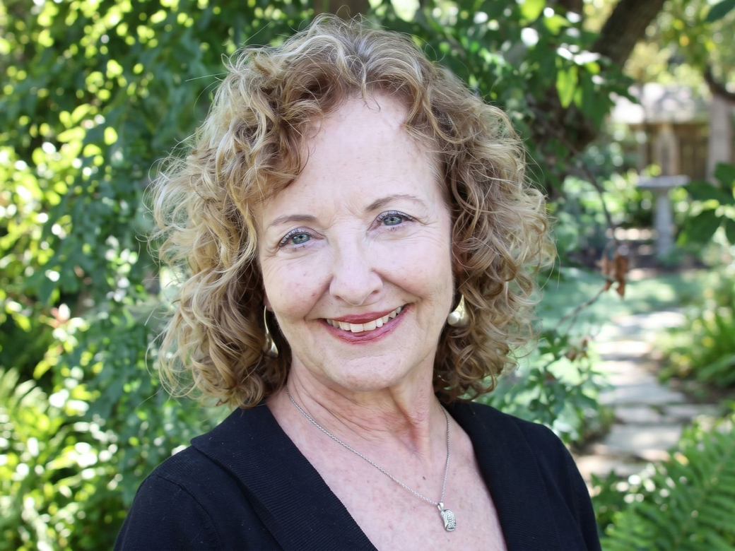 Smiling person outdoors in a garden, with curly hair and a black top.