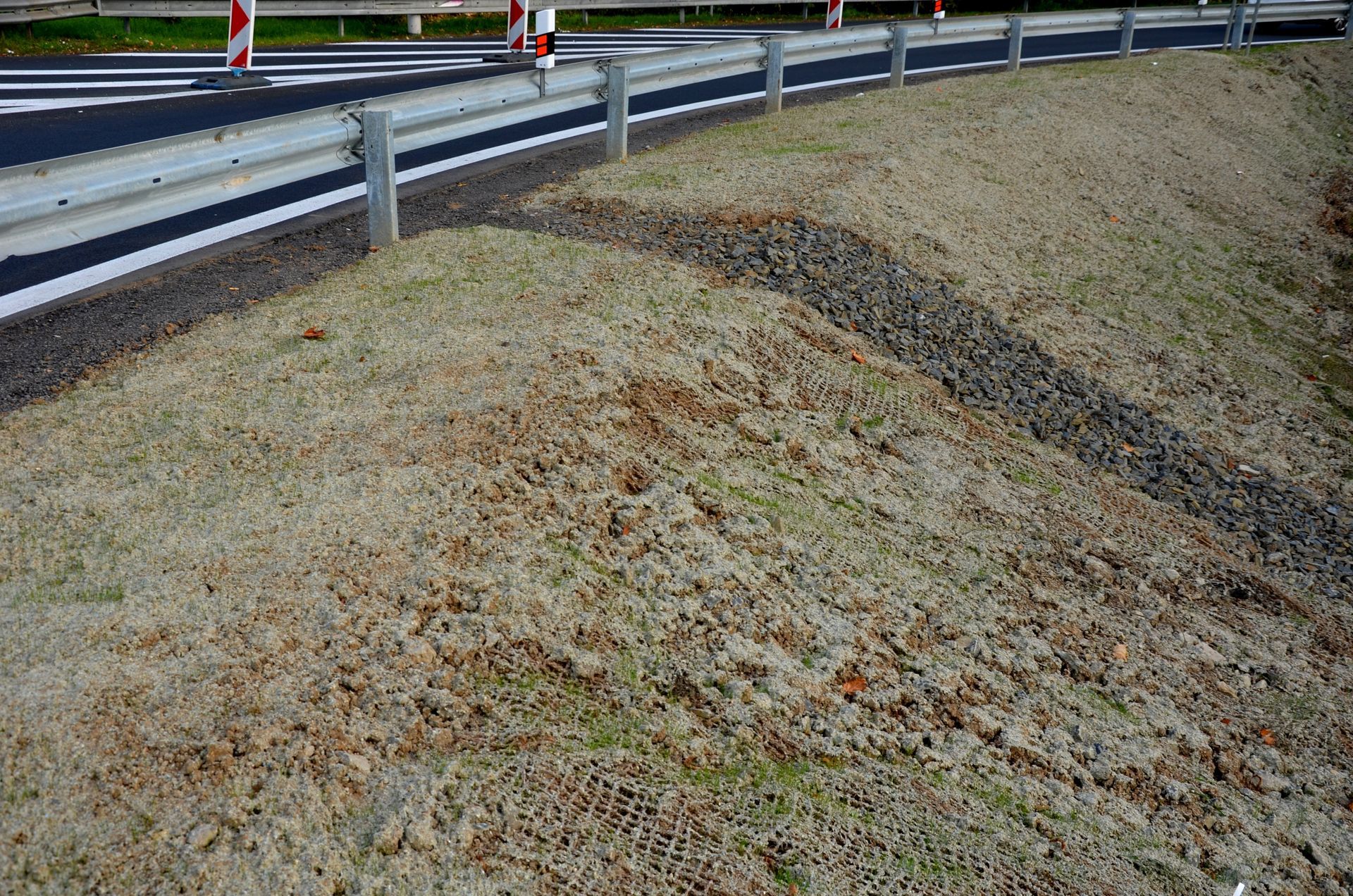 A roadside slope with a gravel drainage trench leading from a guardrail onto a grassy embankment.