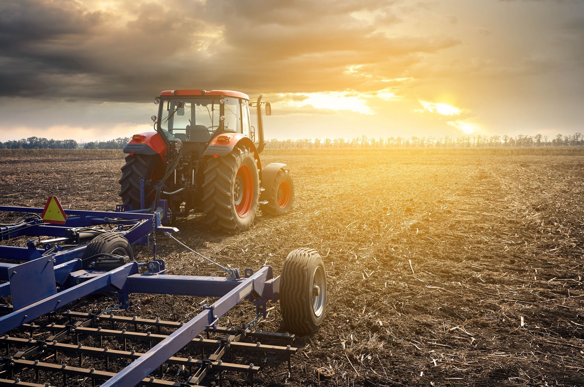 A red tractor pulling farm equipment through a field under a sunset sky with dark, dramatic clouds.