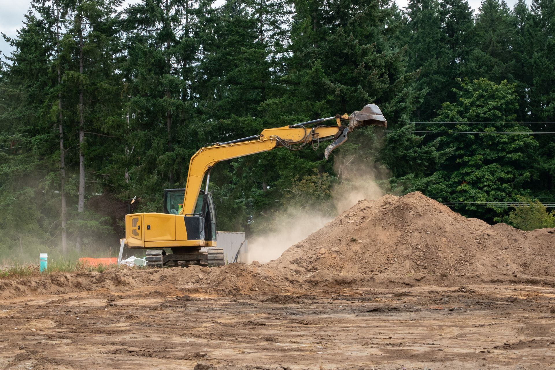 A yellow excavator works at a construction site, lifting a bucket of dirt against a background of tall green trees.