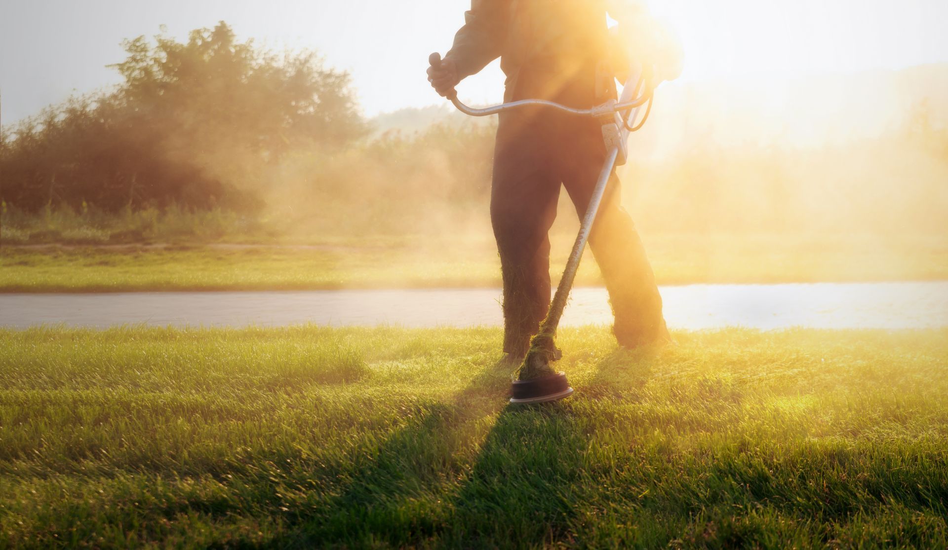 A person uses a string trimmer on a lawn during a sunset, creating a dusty, golden haze.