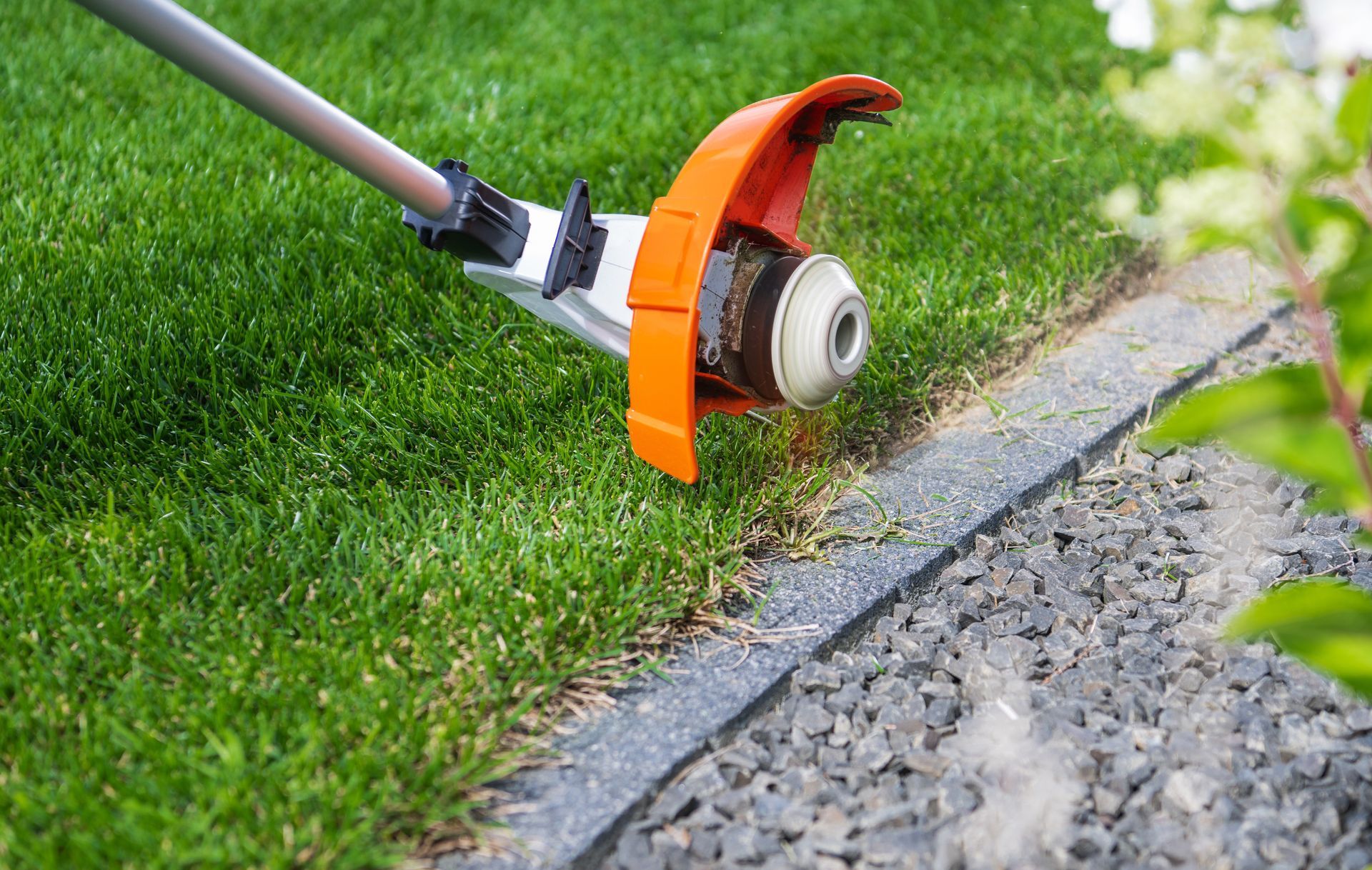 A string trimmer with an orange guard cuts the edge of a green lawn along a stone border.